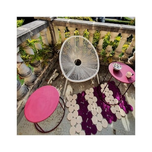 May include: White wicker chair with a round, pink side table and a round, burgundy side table with a white and black patterned bowl on top. The chair and tables are on a patio with a purple and white woven rug.