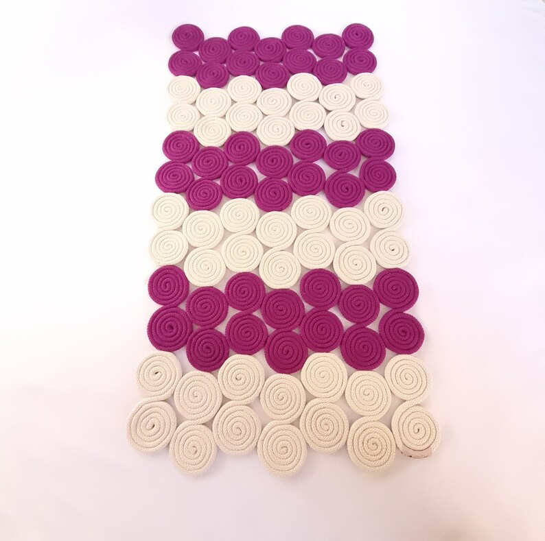 May include: A white and pink woven rug made of coiled rope. The rug is made up of rows of circles, with alternating colours of white and pink.