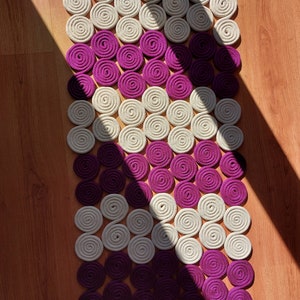 May include: A decorative rug made of woven circles in shades of cream and purple. The circles are arranged in a diagonal pattern, creating a unique and modern design.