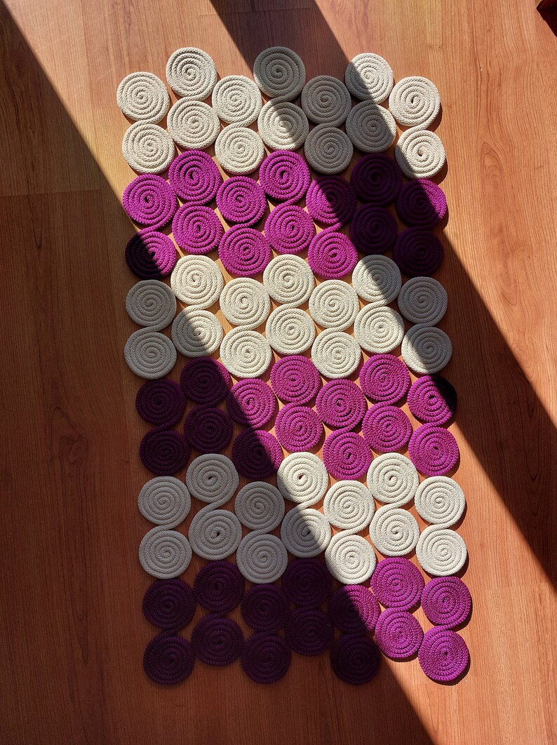 May include: A decorative rug made of woven circles in shades of cream and purple. The circles are arranged in a diagonal pattern, creating a unique and modern design.