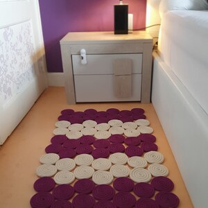 May include: A white and purple round rug made of coiled rope. The rug is on a light orange floor in a bedroom with a white bedside table and a lamp with a floral shade.