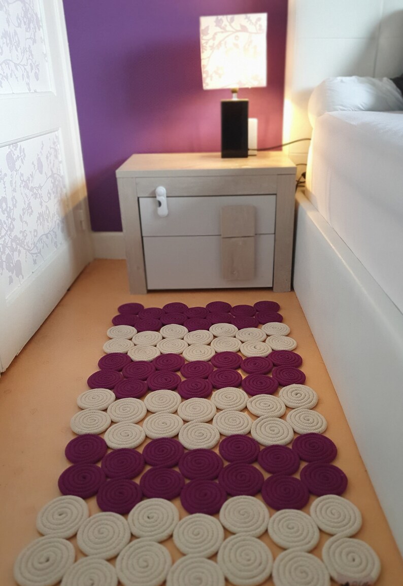 May include: A white and purple round rug made of coiled rope. The rug is on a light orange floor in a bedroom with a white bedside table and a lamp with a floral shade.