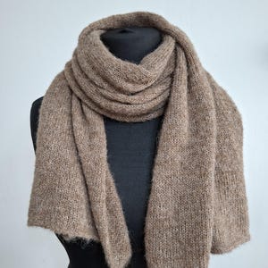 May include: A knitted scarf in a neutral brown color is draped over a black mannequin. The scarf is made of a soft, textured yarn, and the ends have a slightly fringed edge. The scarf is designed to be worn around the neck.