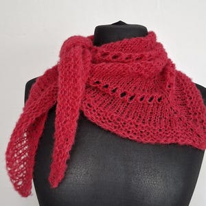Pode incluir: Um xale triangular vermelho vibrante com um delicado padrão de renda aberta e uma aparência felpuda e texturizada. O cachecol está drapeado em um manequim preto, exibindo sua construção leve e arejada e detalhes intrincados e repetitivos.