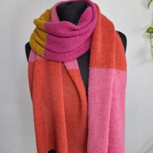 May include: A colorful scarf in shades of pink, orange, and gold. The scarf is draped over a black mannequin, showcasing its rectangular shape and soft texture. The color-blocked design adds a modern touch to this cozy accessory.