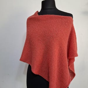 Peut inclure: Un poncho tricoté de couleur corail présenté sur un mannequin noir. Le poncho est drapé sur les épaules, mettant en valeur son tricot texturé et son design fluide. La teinte chaude et l'aspect doux suggèrent un accessoire confortable.