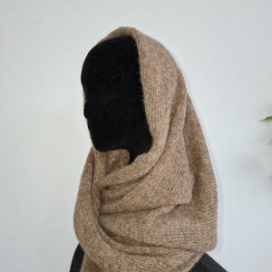 Op de afbeelding: Een gebreide col sjaal in een bruinachtige tint wordt getoond op een zwarte mannequin hoofd. De sjaal, gemaakt van zacht, getextureerd garen, is ontworpen om om de nek en het hoofd te worden gedragen. De neutrale kleur biedt veelzijdigheid.