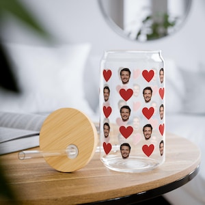 May include: A clear glass can-shaped drinking glass with a bamboo lid and straw. The glass is decorated with red hearts and images of a person's face. The lid is round and made of light-colored wood.