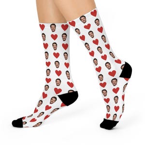 May include: White socks with a repeating pattern of red hearts and a man's face. The socks have black heels and toes. The socks are worn on a person's legs.