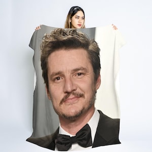 May include: A white blanket featuring a large portrait of a man in a black tuxedo with a black bow tie. The man has brown hair and a mustache. The blanket is held up by a person.
