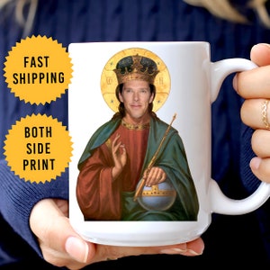 May include: White ceramic mug featuring a portrait of a man in a religious icon style. The image includes a crown, halo, and globe. The mug is held by a person.