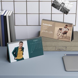 May include: Two spiral-bound desk calendars for the year 2026. One calendar is open to August, featuring a teal background and a photo. The other calendar displays a photo and the text "Calendar 2026 Sebastian Stan". A black and white photo of a bridge is attached to the wall.