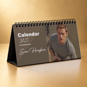 May include: A desk calendar for the year 2027 featuring a spiral binding at the top. The left side has a dark brown background with the white text Calendar 2027 and Sam Heughan. The right side displays a photograph of a person in a gray sweater.