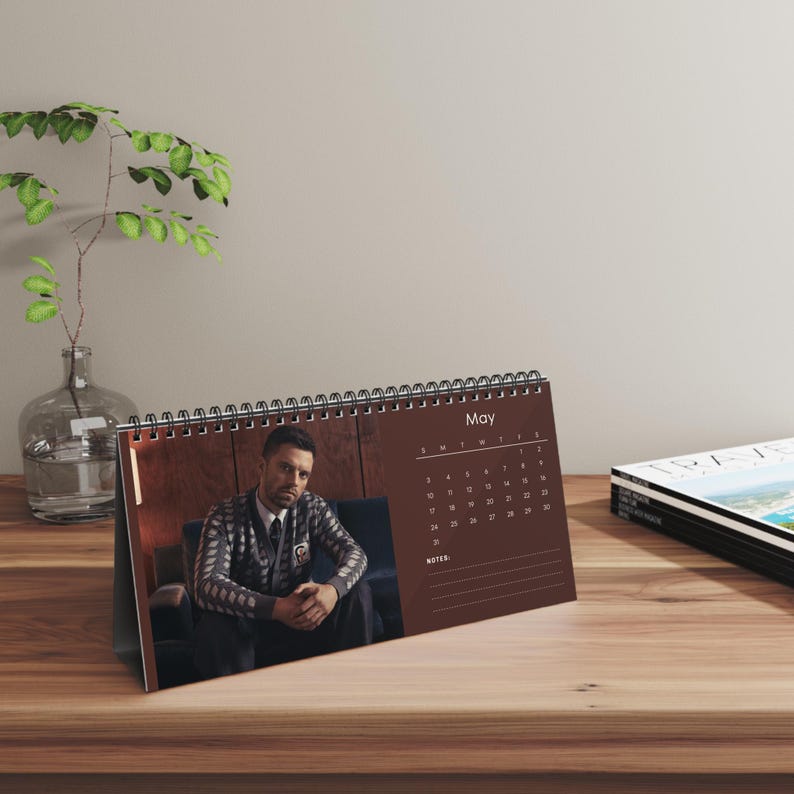 May include: A brown desk calendar for May, featuring a photo of a person in a patterned sweater. The calendar has a spiral binding and a dark brown background. The calendar is on a wooden surface.
