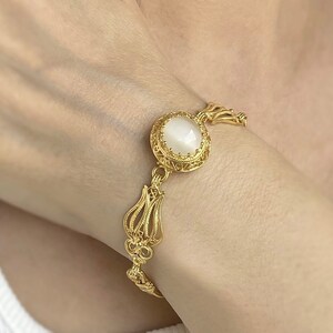 K&ouml;nnte beinhalten: Ein goldfarbenes Armband- und Ohrring-Set mit einem wei&szlig;en Stein in der Mitte jedes St&uuml;cks. Der Schmuck hat ein filigranes Design.