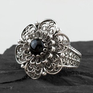 May include: A silver filigree ring with a black gemstone in the center. The ring is set on a black rock.