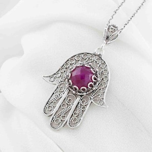 May include: A silver Hamsa hand pendant necklace with a large, round, deep pink gemstone at the centre. The pendant features intricate filigree detailing and hangs on a delicate chain. The necklace rests on a white fabric background.