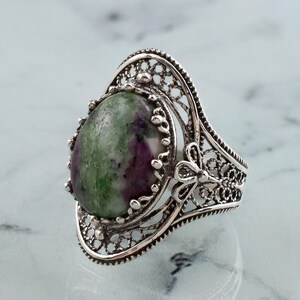 May include: A silver ring with a green and purple gemstone set in a filigree design.