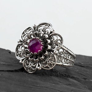 Peut inclure: Une bague en filigrane argentée avec une grande pierre précieuse ronde de couleur violette au centre. La bague est conçue dans un motif floral.