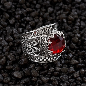 Può includere: Un anello d'argento con un design intricato e una grande pietra rossa.
