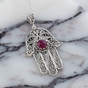 Op de afbeelding: Een zilverkleurige Hamsa hand hanger ketting met een facetgeslepen, ronde, rode edelsteen in het midden. De hanger heeft ingewikkelde filigraan details en hangt aan een delicate ketting. De ketting ligt op een wit en grijs marmeren oppervlak.