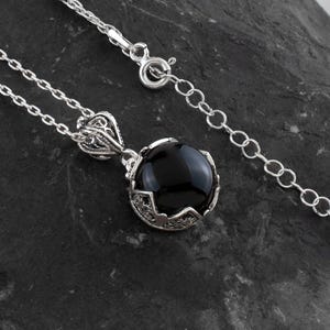 May include: A silver necklace with a round, black gemstone pendant. The pendant is set in a decorative, filigree-style setting. The chain is delicate and has a lobster clasp.