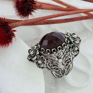 May include: A silver ring with a large, dark red gemstone set in a filigree design. The ring is surrounded by delicate floral accents.