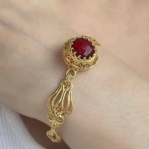 May include: A gold filigree bracelet featuring a large, faceted, red gemstone. The bracelet has a floral design with intricate details and a secure clasp. The bracelet is worn on a wrist.