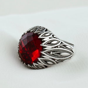 Könnte beinhalten: Ein silberner Ring mit einem verzierten Design, das mit einem großen, roten Edelstein verziert ist.