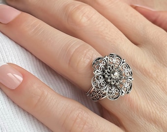 May include: A silver filigree flower ring with intricate openwork design. The ring features a central raised element and detailed petal patterns. The ring is worn on a finger, showcasing the delicate craftsmanship and floral theme.