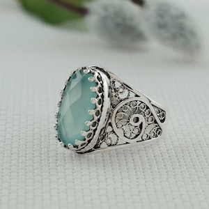 Pode incluir: Um anel de prata com uma pedra preciosa oval azul claro. A pedra é engastada numa banda de prata calada decorativa com um design de filigrana. O anel tem uma estética vintage ou antiga.