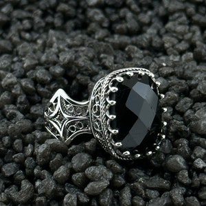 May include: A silver ring with an ornate design and a large black gemstone. The ring is set against a background of small black stones.