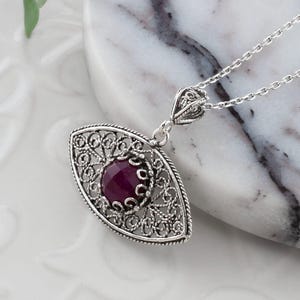 May include: A silver filigree pendant necklace with a deep red gemstone center. The pendant is an oval shape with intricate openwork detailing. The necklace is displayed on a white marble surface, with a red flower in the background.