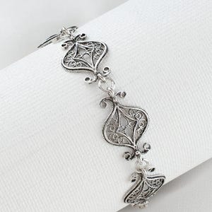 Può includere: Un braccialetto in filigrana d'argento dal design ornato. Il braccialetto presenta tre elementi decorativi simmetrici collegati da piccoli anelli. L'intricato motivo traforato è visibile su uno sfondo bianco. Il braccialetto è lungo circa 18 cm.