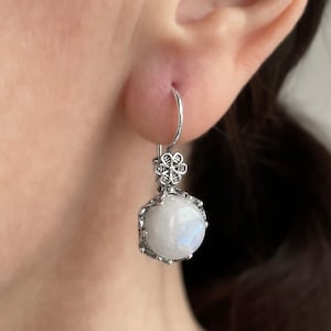 May include: Silver dangle earrings featuring a white moonstone gemstone and a floral design.
