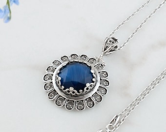 Peut inclure: Un collier en argent avec un grand pendentif rond bleu foncé. Le pendentif est entouré d'un cadre argenté décoratif. Le collier est posé sur une surface blanche, avec des fleurs bleues en arrière-plan. La chaîne est fine et possède un fermoir.