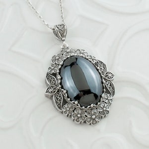 May include: A silver necklace featuring an oval, dark gray gemstone pendant. The pendant is framed by a detailed floral design. The necklace chain is delicate and silver, with a decorative bail.