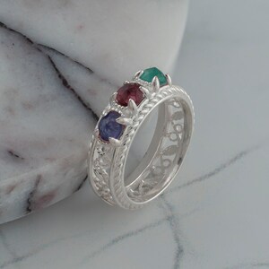 May include: A silver ring with a delicate filigree design and four gemstones. The gemstones are green, red, and blue.