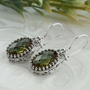 May include: A pair of silver drop earrings featuring oval-shaped, faceted olive-green gemstones. The stones are set in ornate, scalloped silver bezels with intricate metalwork detailing along the sides and attached to simple silver ear wires.