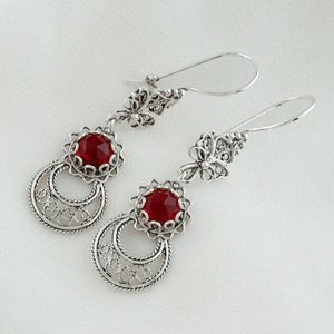 May include: A pair of silver dangle earrings with red gemstone accents. The earrings feature intricate filigree designs and a crescent moon shape.