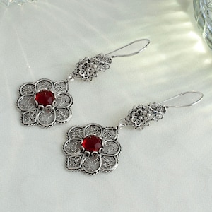 Può includere: Un paio di orecchini in filigrana d'argento con accenti di pietre preziose rosse. Gli orecchini presentano un delicato design floreale e pendono da un gancio.