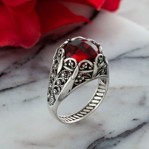 May include: A silver ring with a large, red gemstone set in a filigree design. The ring is on a white marble surface.