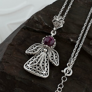 Könnte beinhalten: Eine silberne Filigran-Engel-Anhänger-Kette mit einem violetten Edelstein. Die Kette hat eine zarte Kette und einen floralen Verschluss.