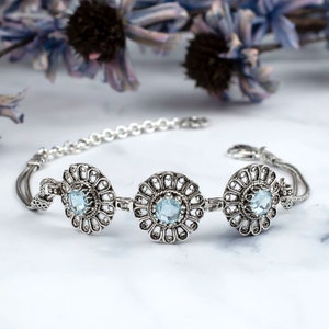 May include: A silver bracelet with three round, filigree-style charms, each featuring a light blue gemstone in the center.