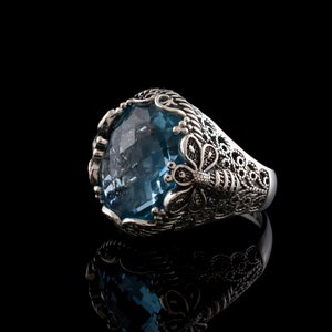 Può includere: Un anello d'argento con una grande pietra preziosa blu e dettagli filigranati intricati. L'anello presenta un design a farfalla sul lato.