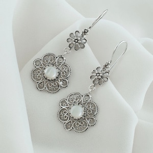Op de afbeelding: Een paar zilveren filigraan oorbellen met witte parelmoer stenen. De oorbellen hebben een delicaat bloemmotief en hangen aan een haak.