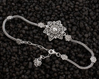 May include: A silver chain bracelet with a large, intricate flower charm and smaller flower accents. The bracelet has a lobster clasp closure.