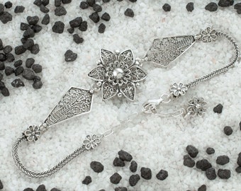 May include: A silver filigree bracelet with a large flower-shaped charm in the center. The bracelet is made of a delicate chain and has smaller flower charms on either side of the main charm.