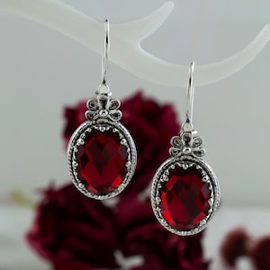 May include: A pair of silver dangle earrings with red gemstone drops. The earrings feature a floral design at the top and a detailed oval frame around the gemstone.