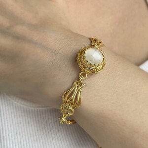 K&ouml;nnte beinhalten: Ein goldenes Armband mit einem wei&szlig;en ovalen Stein, der in einem filigranen Design gefasst ist. Das Armband hat einen tulpenf&ouml;rmigen Anh&auml;nger.